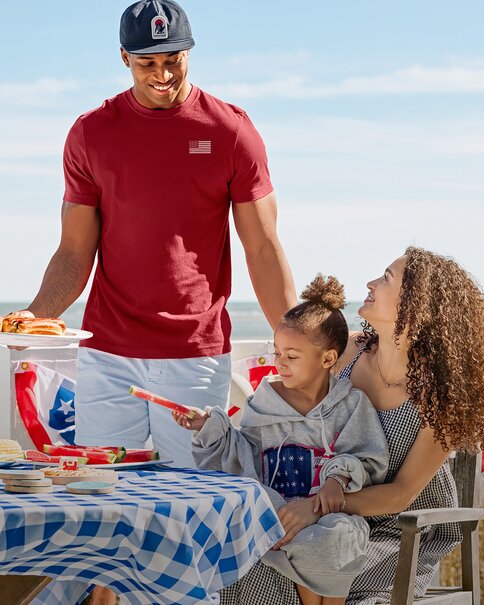 A family enjoys a sunny beachside picnic around a blue gingham-covered table with watermelon slices and hot dogs. A man in a red T-shirt stands beside a seated woman and child, creating a relaxed patriotic summer scene with the ocean in the background.