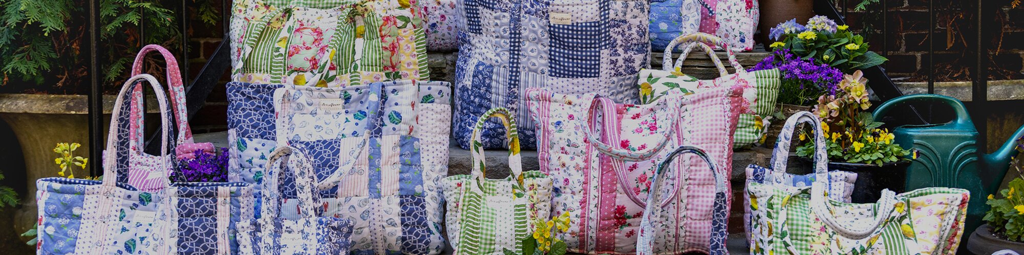 Colorful handmade quilted Strawflower tote bags and cushions in floral and patchwork patterns displayed outdoors on steps, surrounded by potted flowers and a watering can.