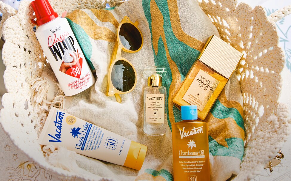 A woven crochet beach bag holds an assortment of summer skincare and beach essentials from Vacation, including sunscreen, tanning oil, sunglasses, and fragrance bottles layered over a striped towel. Warm sunlight highlights the neutral textures and golden tones, creating a relaxed, vintage-inspired vacation aesthetic.