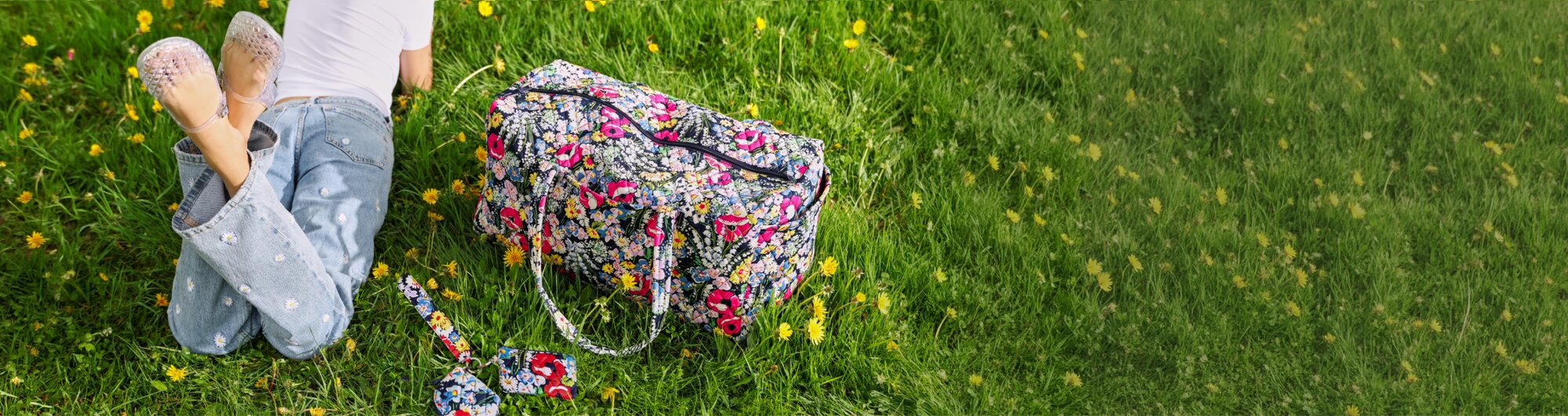 Person lying on green grass dotted with yellow flowers, wearing a white T-shirt, light-wash jeans with embroidered daisies, and clear shoes. A large Vera Bradley duffel bag in Poppy Fields and matching keychain pouch rest beside them, creating a colorful spring-inspired outdoor scene.