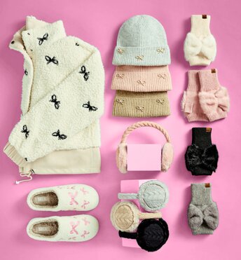 A holiday bow sweater, three knit beanies, cozy slippers, ear muffs, and head bands laying on a pink background