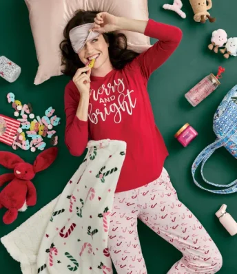 Image of a woman laying on a green background wearing holiday pajamas surrounded by festive items