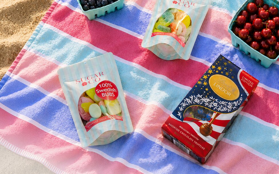 A red, white, and blue striped beach towel topped with assorted Swedish candy, chocolate, and fresh berries arranged for a summer picnic.