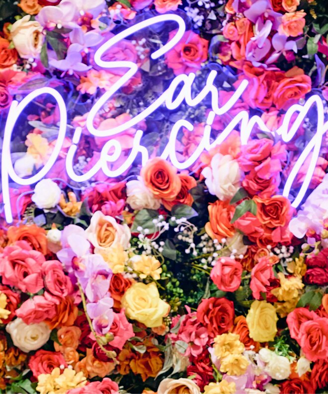 Colorful floral wall with a glowing neon “Ear Piercing” sign.