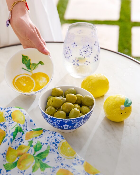 A Mediterranean-inspired tabletop display features bowls of green olives, fresh lemons, and a patterned blue-and-white drinking glass on a marble table. A hand reaches toward a ceramic bowl decorated with lemon illustrations, while a lemon-print cloth napkin drapes across the foreground. Bright sunlight and greenery create a fresh, summery atmosphere.