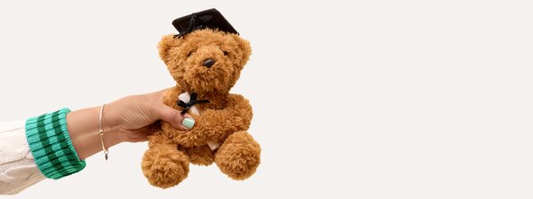 A hand in a cream sweater with green striped cuffs holds a plush teddy bear dressed in a graduation cap and bow tie. The stuffed animal is photographed against a simple neutral background, creating a cheerful graduation gift-inspired scene.