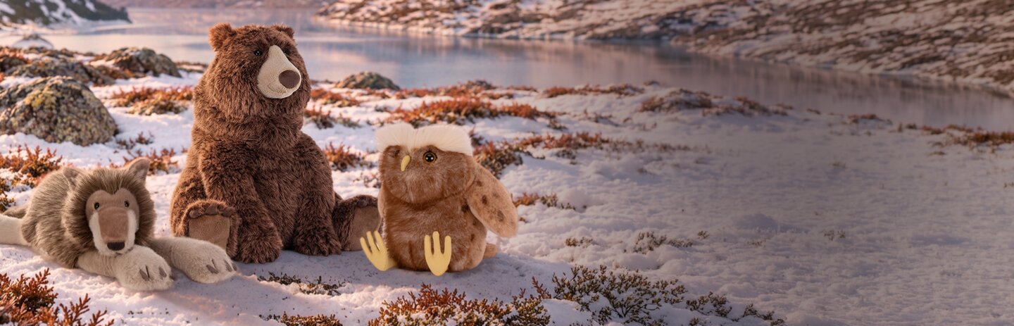 Three plush woodland Jellycats—a wolf, brown bear, and owl—sit together in a snowy tundra landscape with mountains in the background.