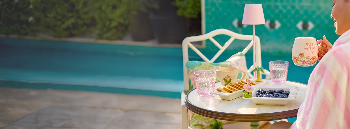 A woman in a pink and white striped robe sits by a pool, holding a Strawflower mug that says “Best Mom Ever,” next to a small table set with Nora Fleming trays holding blueberries and cookies, and decorative glasses in a sunny poolside setting.