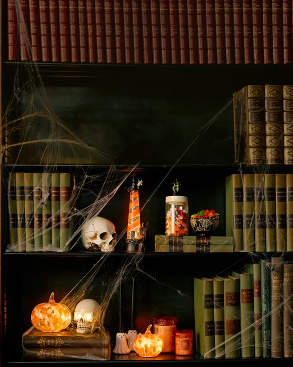 A bookshelf decorated with glowing pumpkins, skull accents, candles, cobwebs, and vintage books creates a cozy Halloween-inspired home décor display.
