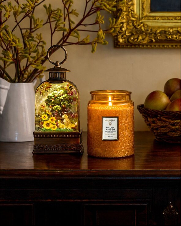A lit amber candle and illuminated decorative lantern are displayed on a wooden table with a basket of apples and seasonal greenery in the background.