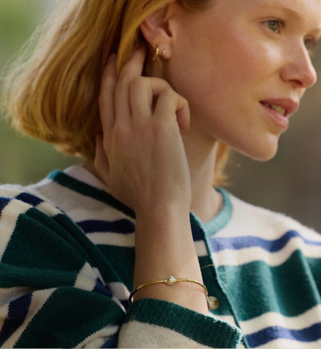 Close-up of a woman wearing a gold Cape Cod bracelet with a pearl accent and small hoop earrings.