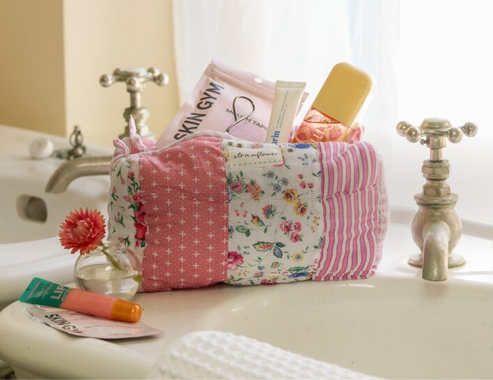 Pink-and-floral patchwork quilted cosmetic bag filled with beauty products, displayed beside a bathroom sink. 
