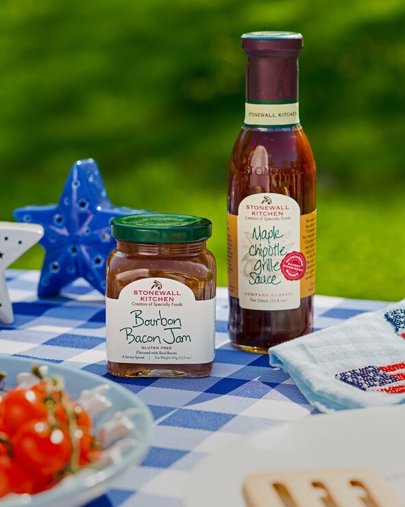 Stonewall Kitchen Bourbon Bacon Jam and Maple Chipotle Grille Sauce displayed on a blue gingham picnic table outdoors.