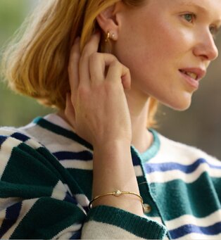 Close-up of a woman wearing a gold Cape Cod bracelet with a pearl accent and small hoop earrings.