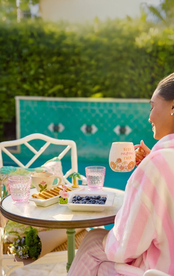 A pregnant woman lounges on a lounge chair outdoors, smiling and wearing sunglasses and a pink floral pajamas. She holds a lip gloss in one hand and a Strawflower cosmetic bag in the other. A pink-and-orange towel covers the chair, and a large Strawflower tote sits nearby. Green bushes and plants fill the background, creating a relaxed, sunny poolside setting.