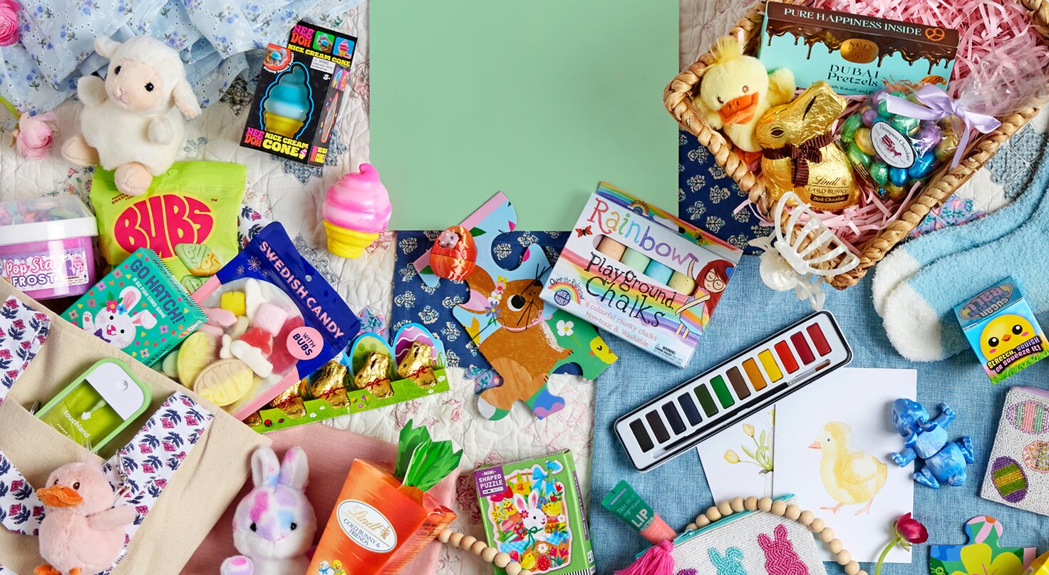A colorful flat lay of Easter basket goodies arranged on pastel fabrics and quilts, featuring Palm Pals plush bunny and duck toys, candy like BUBS gummies and Swedish candy, Lindt chocolate bunnies, a duck-themed puzzle, watercolor paint set, rainbow playground chalk, small toys like NeeDoh and DopeSlimes, Tree Hut lip balm and scrub, Touchland hand sanitizers, and a woven basket filled with treats and a plush chick.