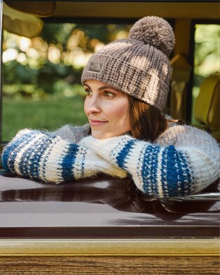 Woman wearing a gray pom-pom beanie and blue-and-white patterned sweater, leaning out of a car window.