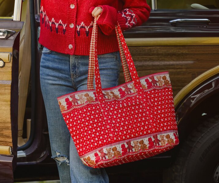 Woman in a red cardigan and jeans carrying a red holiday-print Vera Bradley tote bag beside a vintage car.