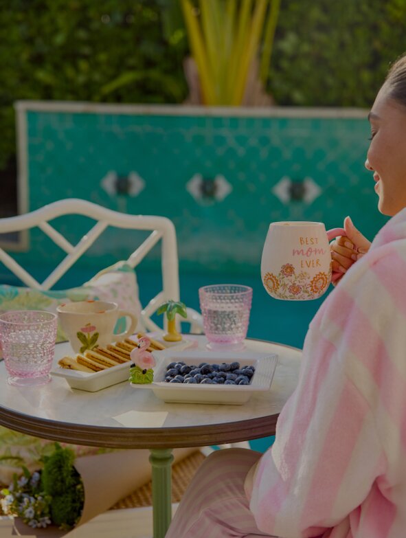 A woman in a pink and white striped robe sits by a pool, holding a Strawflower mug that says “Best Mom Ever,” next to a small table set with Nora Fleming trays holding blueberries and cookies, and decorative glasses in a sunny poolside setting.