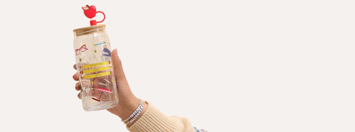 A hand wearing beaded bracelets holds a ceramic mug with a yellow ruler-shaped handle and the phrase “Teaching is a Work of Heart” beside a raised red heart design.