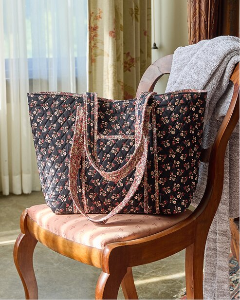 Black floral quilted Vera Bradley tote bag with patterned trim and double shoulder straps, displayed on a wooden chair beside a cozy gray blanket.