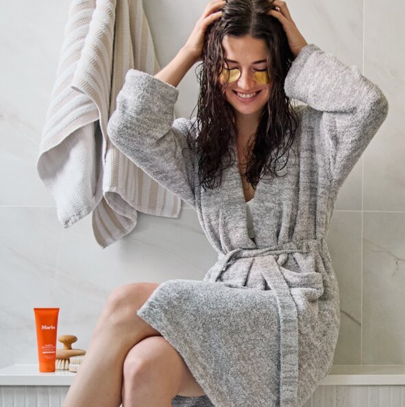 Model in a bathroom wearing a gray robe and under eye masks, with Marin skincare next to her