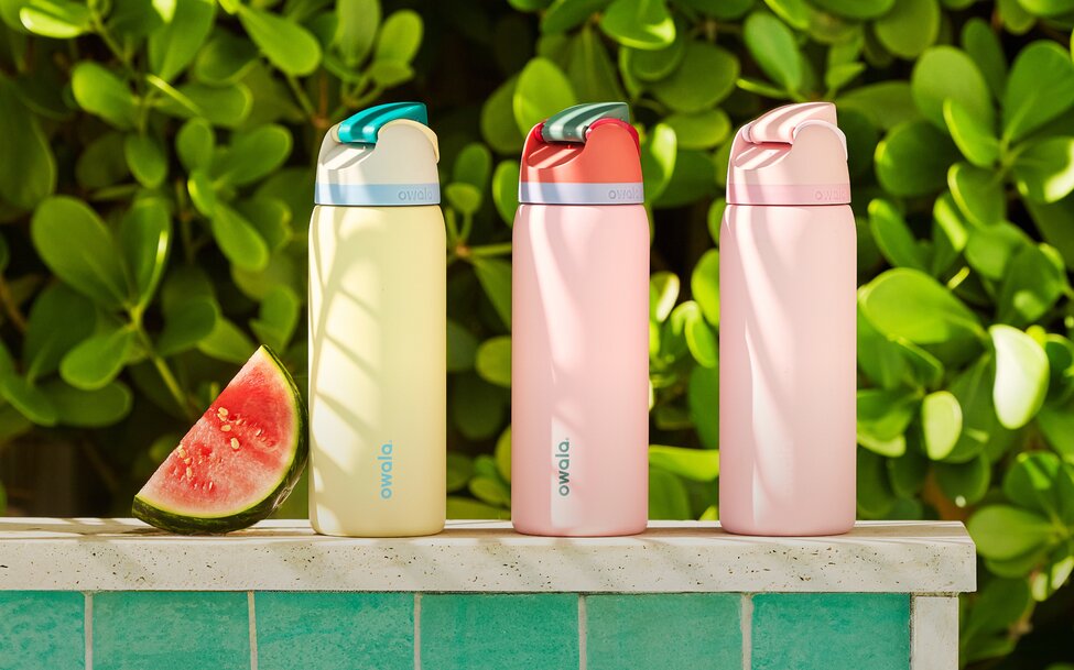 Three pastel-colored Owala insulated water bottles are displayed on a turquoise tiled ledge beside a slice of watermelon, with lush green tropical foliage in the background. Bright sunlight and playful color combinations create a fresh, summery poolside aesthetic.