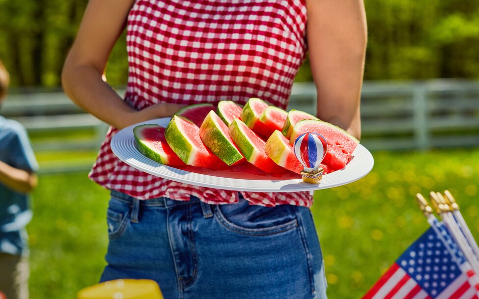 Person in a red gingham top holding a platter of sliced watermelon at an outdoor summer picnic with patriotic Nora Fleming mini ornament