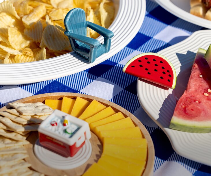 A family enjoys a sunny beachside picnic around a blue gingham-covered table with watermelon slices and hot dogs. A man in a red T-shirt stands beside a seated woman and child, creating a relaxed patriotic summer scene with the ocean in the background.