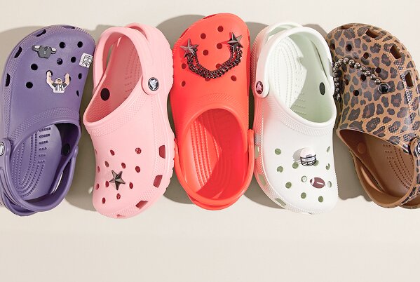 A row of five colorful Crocs clogs arranged side by side on a light background. Each shoe features decorative charms: a purple pair with small icons, a pink pair with a star, a bright red pair with star and chain accents, a pale green pair with simple charms, and a brown pair with a leopard print pattern.