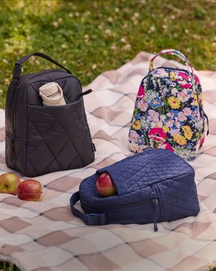 Vera Bradley quilted lunch bags and a floral backpack arranged on a picnic blanket with apples.