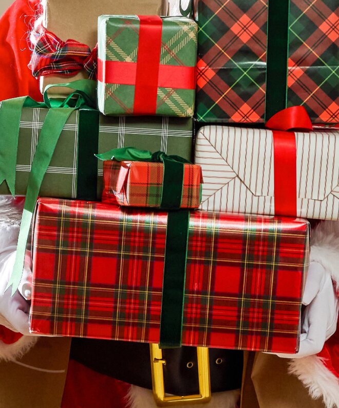 Santa Claus holding a stack of Christmas presents wrapped in festive red and green plaid paper with coordinating ribbons.