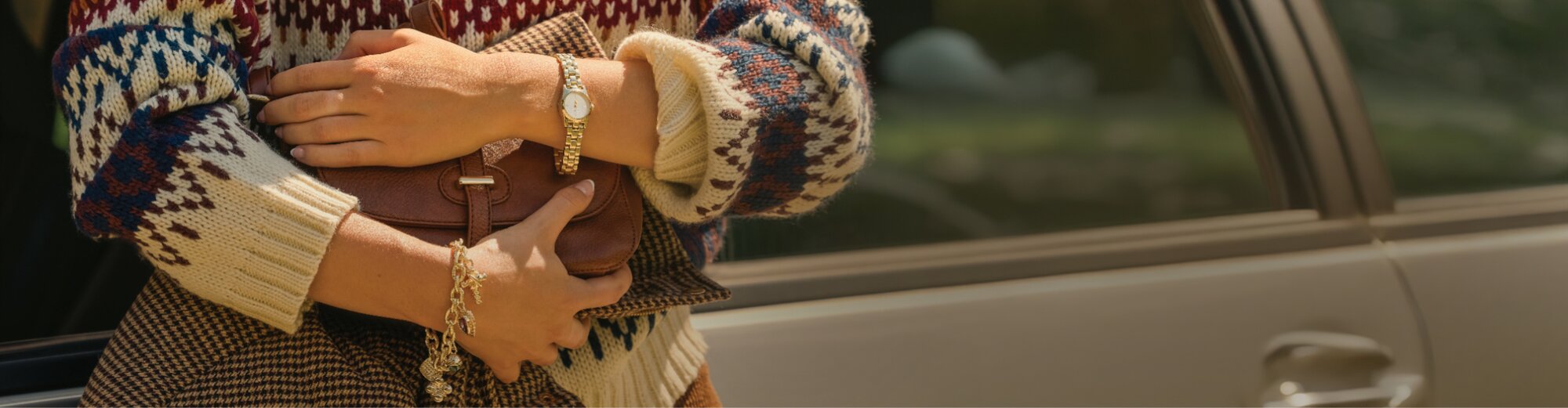 Person in a patterned sweater holding a brown handbag beside a car.