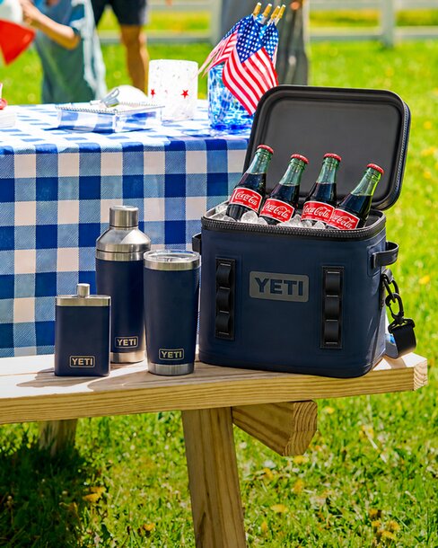 Navy YETI cooler filled with glass Coca-Cola bottles alongside matching drinkware at a backyard picnic setup with American flags.