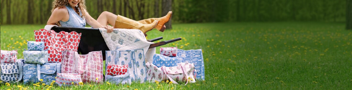 Woman sitting on grass behind a collection of floral and patterned Strawflower tote bags, duffels and cosmetic bags.