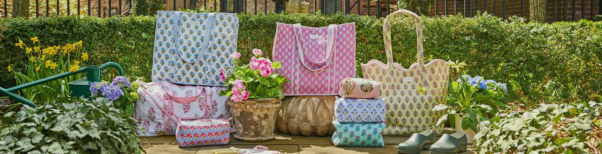 Assortment of Strawflower patchwork quilted tote bags arranged in a sunlit garden, surrounded by blooming flowers, greenery, and a pair of green garden clogs on a stone patio.