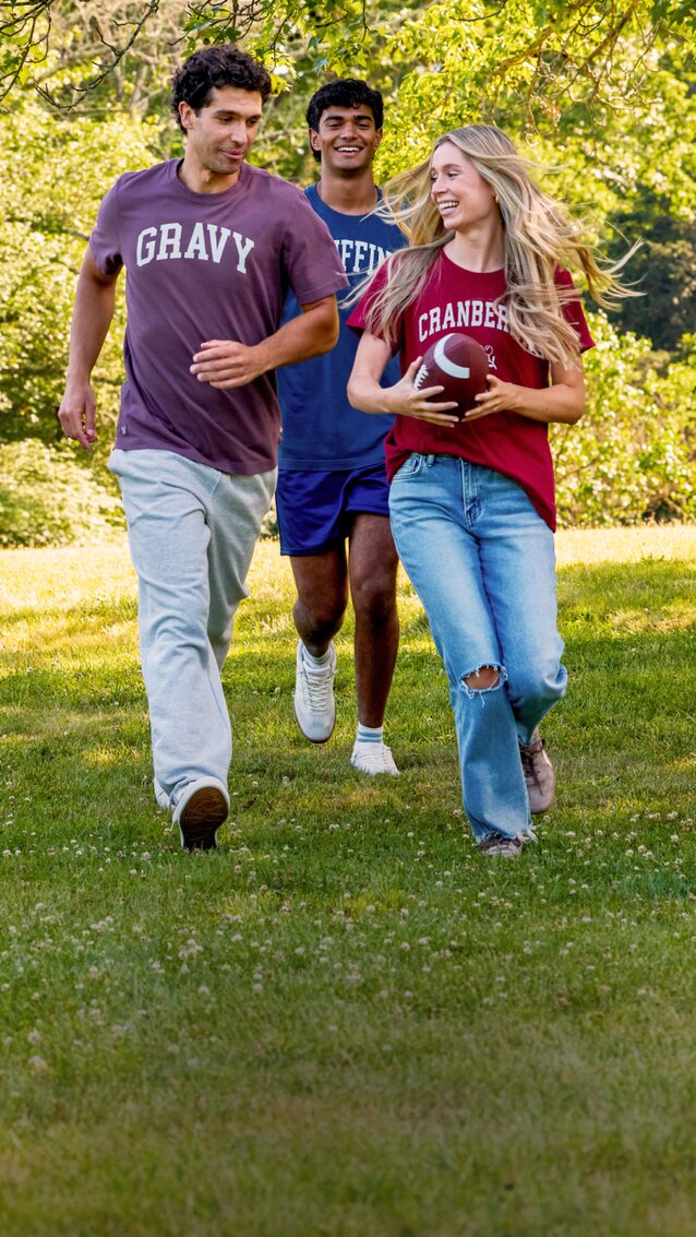Five friends running across a grassy park in casual Thanksgiving-themed graphic tees, with one holding a football.