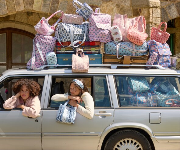 A car loaded with colorful quilted backpacks, totes, duffel bags, and luggage is parked outside a stone building while two people lean out the windows.