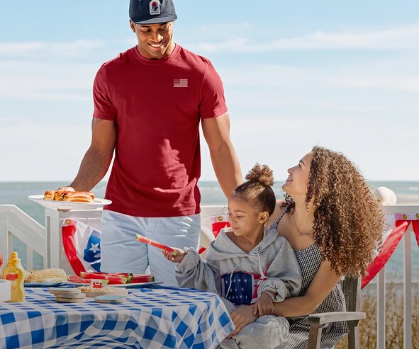 A family enjoys a sunny beachside picnic around a blue gingham-covered table with watermelon slices and hot dogs. A man in a red T-shirt stands beside a seated woman and child, creating a relaxed patriotic summer scene with the ocean in the background.