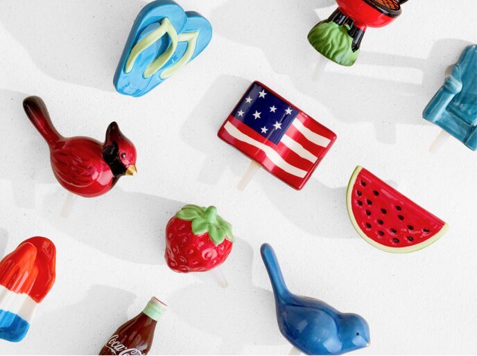 Image of various Nora Fleming mini ornaments, featuring blue flip flops, an American flag, a watermelon slice, a cardinal, a strawberry, a blue bird, a red white and blue popsicle, and a bottle of Coca Cola.