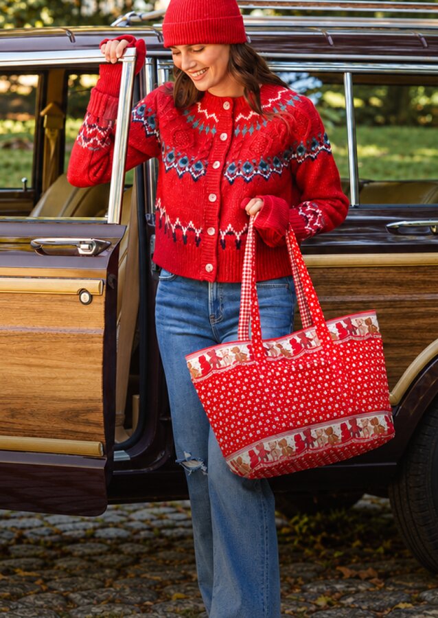Woman in a red sweater and jeans carrying a red holiday-print Vera Bradley tote bag beside a vintage wood-paneled car.