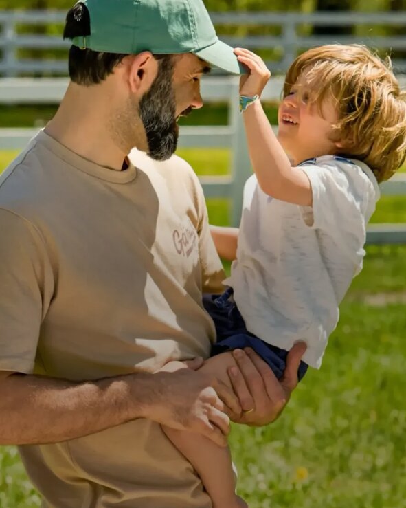 Father and child outdoors in a sunny grassy field, with the child reaching up to touch the man’s green baseball cap near a white fence.