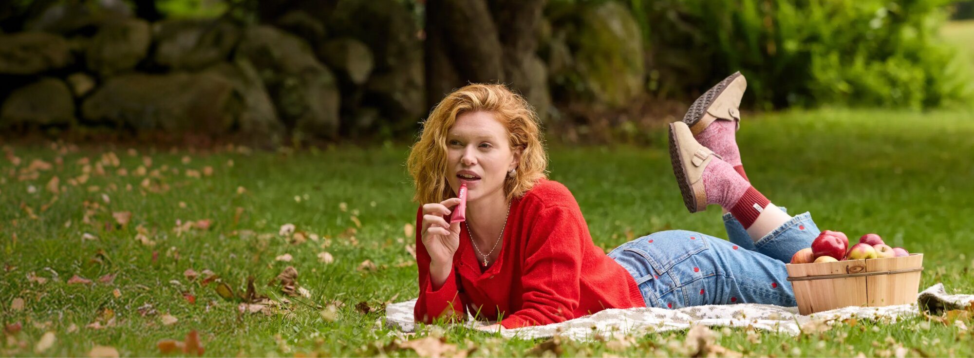 Woman in a red sweater relaxing on a picnic blanket beside a basket of apples.