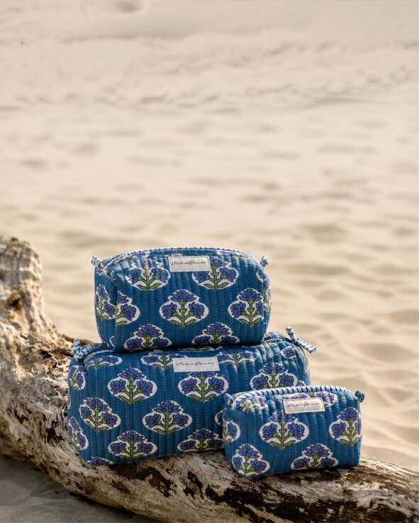 A set of three Strawflower blue floral quilted pouches displayed on driftwood in the sand.