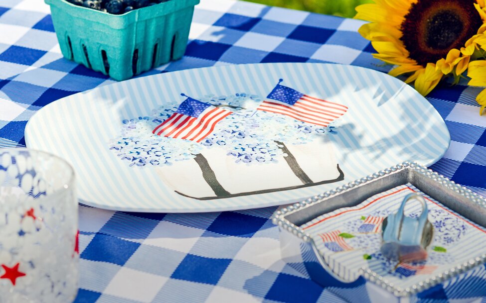 Patriotic outdoor picnic table setting featuring blue-and-white gingham linens, decorative serving pieces, fresh blueberries, and sunflower accents.