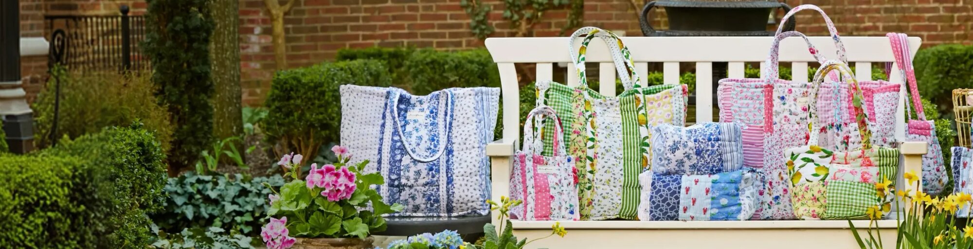 Colorful handmade quilted Strawflower tote bags and cushions in floral and patchwork patterns displayed outdoors on a bench, surrounded by potted flowers and a watering can.