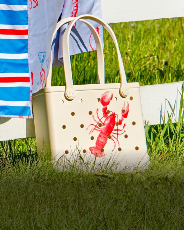 White Bogg tote bag with a red lobster design sitting in the grass beside patriotic summer décor.