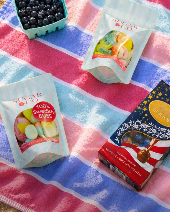 A red, white, and blue striped beach towel topped with assorted Swedish candy, chocolate, and fresh berries arranged for a summer picnic.