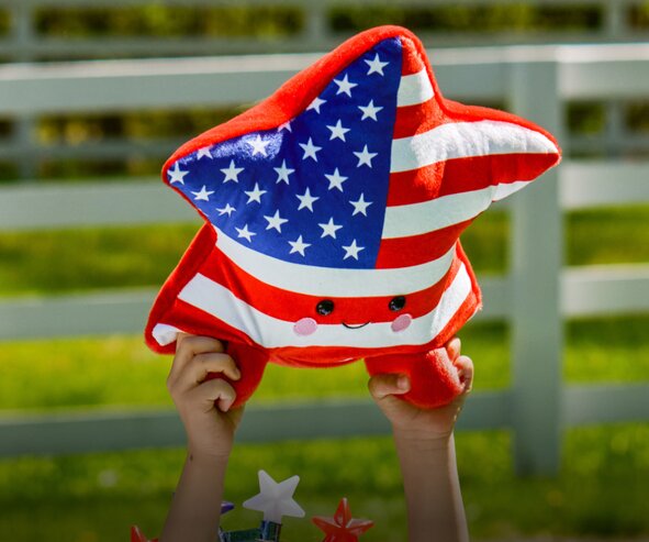 Child holding a red, white, and blue star-shaped plush pillow in front of a white fence outdoors.