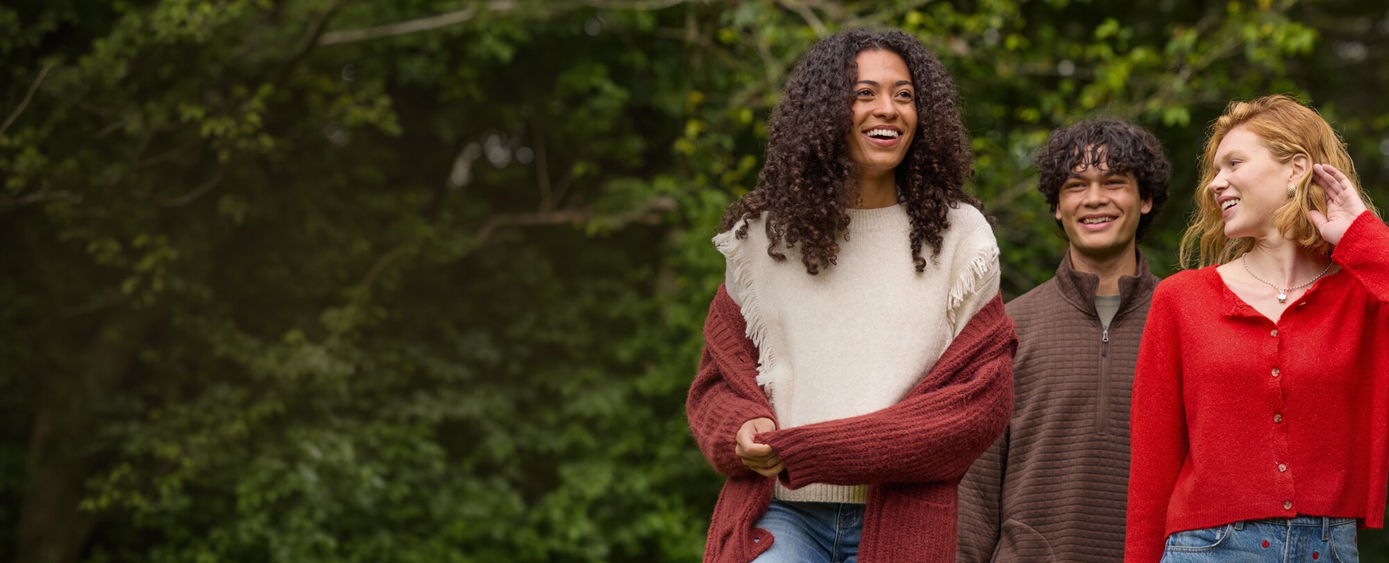 Three friends wearing cozy fall layers smile together outdoors against a leafy green backdrop.